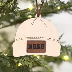 May include: A wooden Christmas ornament shaped like a beanie hat, hanging from a brown leather cord. The hat is light-colored with a brown rectangular patch that reads "NOAH". The ornament is hanging on a Christmas tree.
