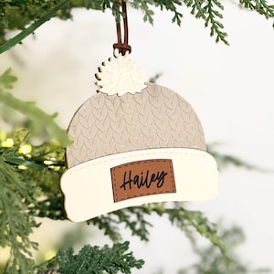 May include: A wooden Christmas ornament shaped like a winter hat, hanging from a brown string. The hat is beige with a white brim and a pom-pom. The name "Hailey" is printed on a brown leather-like patch.