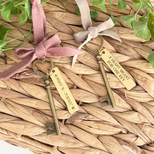 May include: Two vintage-style key favors with attached tags and ribbons. One key has a heart-shaped top, the other has a rectangular top. The tags read "A HEART MEANT FOR ONE KEY" and "41.9265° N -71.8765° W". The keys are on a woven basket.