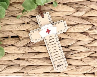Può includere: Un ornamento a forma di croce in legno con un cappello da infermiera e una croce rossa. La croce ha un bordo smerlato e un cordino in pelle per appenderla. Il testo sulla croce recita: "Signore, guida le mie mani e il mio cuore mentre mi prendo cura degli altri. Dammi pazienza, compassione e forza ogni giorno. Amen."