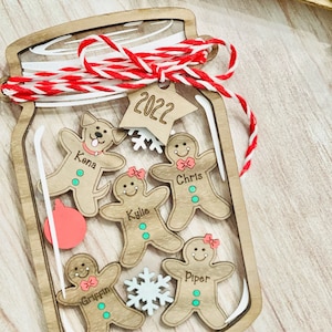 May include: A wooden Christmas ornament shaped like a mason jar with a red and white twine bow. Inside the jar are five gingerbread people with names: Kona, Chris, Kylie, Griffin, and Piper. A star-shaped tag with the year 2022 is attached to the jar.