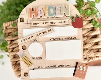 Puede incluir: Un tablero escolar de madera clara con el nombre HAYDEN en letras pastel. Incluye campos de texto para el primer y último día de escuela, edad, cosas favoritas y metas, decorado con un lápiz, una manzana y un cuaderno de notas.