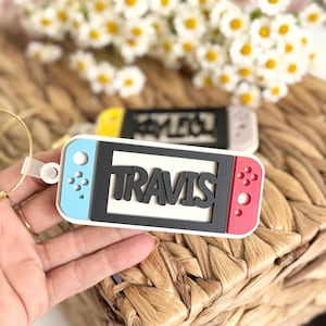 May include: A white, blue, and red keychain shaped like a game console with the name "TRAVIS" in black letters. The keychain has a clear loop and is set against a woven background with white flowers.