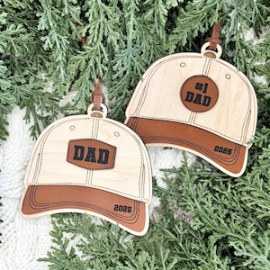 May include: Two wooden baseball cap ornaments with brown leather visors and accents. One ornament reads "#1 DAD" and the other reads "DAD". Both have the year "2025" on the visor. Each ornament has a brown leather loop for hanging.