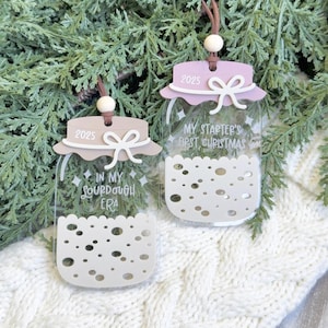 May include: Two clear acrylic jar-shaped Christmas ornaments. Each ornament has a light brown and pink lid with the year 2025. One ornament reads "In my sourdough era" and the other reads "My starter's first Christmas". Both have a white bow.