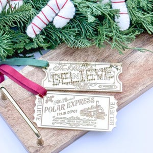 May include: Two gold-colored ticket ornaments with a Christmas theme. The top ornament says "The Polar Express Round Trip" and the bottom ornament says "All Aboard! Polar Express Train Depot Admit One - Round Trip - North Pole" with a train illustration. Both ornaments have the word "Believe" on them.