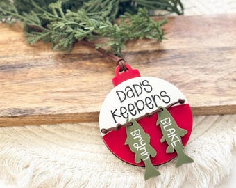 May include: A red and white Christmas ornament shaped like a fishing bobber with the text "Dad's Keepers" on the white side. Two green fish ornaments hang from the bobber with the names "Blake" and "Brynn" written on them.