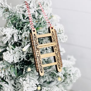 May include: Wooden Christmas ornament shaped like a ladder with the text "The Rose Suchank Ladder Company" on the side. The ornament is hanging on a snowy Christmas tree.