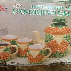 May include: A ceramic tea set with a pineapple design. The set includes a teapot with a lid and four mugs. The teapot and mugs are orange, green, and cream-coloured, with a textured pineapple pattern. The box reads "5 PCS-CONJUNTO PARA TE".