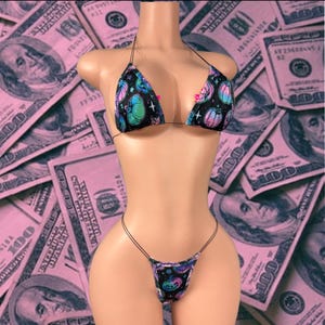 Könnte beinhalten: Ein zweiteiliges Bikini-Set mit schwarzer Stoffbasis und einem sich wiederholenden Muster aus bunten Neon-Kürbissen und kleinen Sternen. Das Oberteil besteht aus zwei dreieckigen Cups mit dünnen schwarzen Trägern und das Unterteil ist ein Tanga.