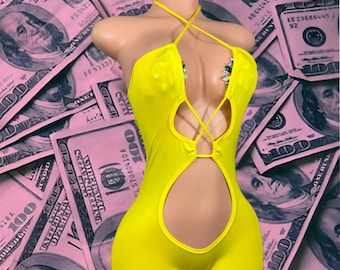 Puede incluir: Un mono amarillo brillante con cuello halter y un corte de ojo de cerradura. La prenda presenta correas entrecruzadas y se muestra en un maniquí. El fondo es un collage de billetes de 100 dólares teñidos de rosa.