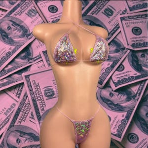 May include: A pink sequined bikini set displayed on a mannequin. The bikini top features a halter neck design and the bottoms are a thong style. The background is a pink-tinted collage of US $100 bills.