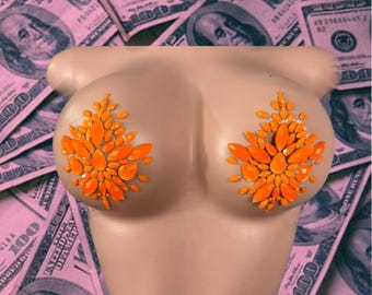 May include: A pair of orange rhinestone nipple pasties arranged in a teardrop-shaped cluster on a beige mannequin torso. The decorative adhesive covers feature faceted orange gems in various sizes, creating a symmetrical floral-like pattern.