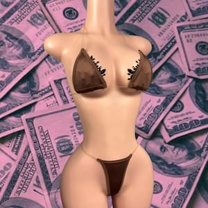 May include: A brown bikini with clear straps and black embellishments on the top cups. The bikini is displayed on a mannequin against a background of pink-tinted hundred-dollar bills.