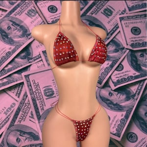 May include: A red bikini set featuring a triangle top and a thong bottom, both adorned with numerous small, clear, reflective rhinestones. The bikini is displayed on a mannequin, showcasing thin red string ties for both the top and bottom pieces.