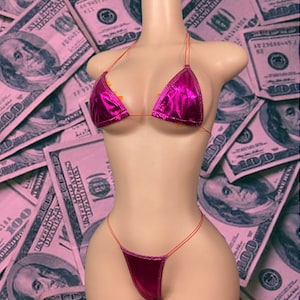 May include: A metallic fuchsia bikini set with a triangle top and string bottom. The bikini is displayed on a mannequin against a backdrop of pink-tinted hundred-dollar bills.