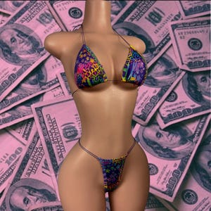May include: A brightly coloured bikini set displayed on a mannequin. The top and bottom feature a vibrant mix of patterns, including leopard print and stars, with shades of pink, yellow, purple, and blue. The background is a pink-tinted image of US $100 bills.