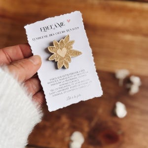 May include: A wooden Edelweiss flower pin with a heart-shaped center is displayed on a white card. The card has text in French, including "FDELAME" and "L'EMBLEME DES COEURS TENACES". The pin is held by a person.