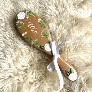May include: Wooden baby brush with a floral design and the name "Mia" in white script. The brush features white and green painted flowers and leaves, with a white ribbon tied at the handle.