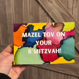 May include: A yellow card with colourful abstract paint splatter in the background. The text "Mazel Tov on your B'mitzvah!" is written in white.