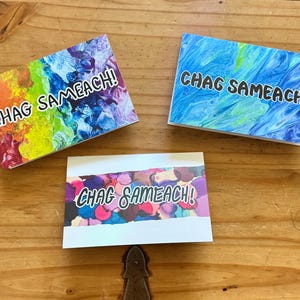 May include: Three greeting cards with the text "CHAG SAMEACH!" on a wooden surface. The cards feature colorful abstract designs in shades of blue, purple, pink, yellow, and green. The cards are rectangular and have white borders.