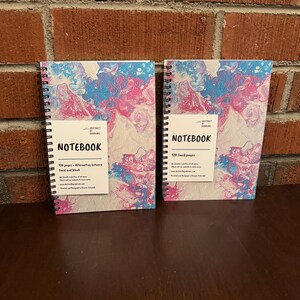 May include: Two spiral bound notebooks with abstract pink and blue marbled covers. The covers feature the text "ABSTRACT BY KARAM" and "NOTEBOOK". The text on the left notebook reads "120 pages - Alternating between lined and blank". The text on the right notebook reads "120 lined pages".