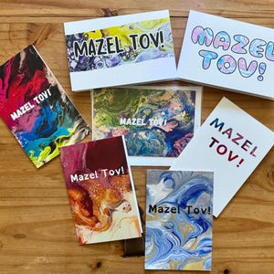 Peut inclure: Plusieurs cartes de vœux avec l'inscription "Mazel Tov!" dans différents styles et couleurs. Les cartes présentent des motifs d'art abstrait aux couleurs vives de rouge, bleu, jaune et vert. Elles sont disposées sur une surface en bois.
