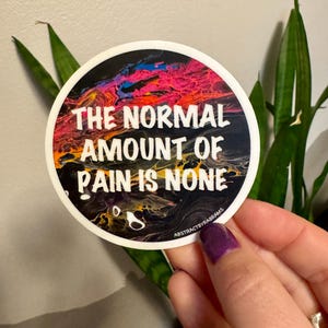 May include: Round sticker with a black, yellow, pink, and blue abstract paint design. The text "THE NORMAL AMOUNT OF PAIN IS NONE" is written in white on the sticker. The sticker is held by a hand with purple nail polish.