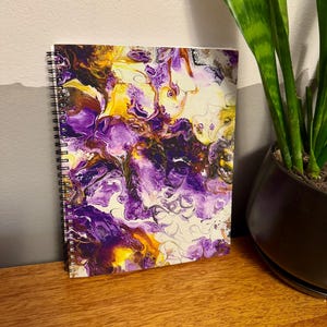 May include: A spiral-bound notebook with a vibrant abstract design. The cover features a swirling pattern of purple, yellow, and white acrylic paint. The notebook is resting on a wooden surface, with a plant in the background.
