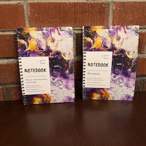 May include: Two spiral bound notebooks with abstract purple, yellow and white marble designs. The covers feature the text "ABSTRACT BY BARBANS" and "NOTEBOOK". Each notebook has 120 lined pages.