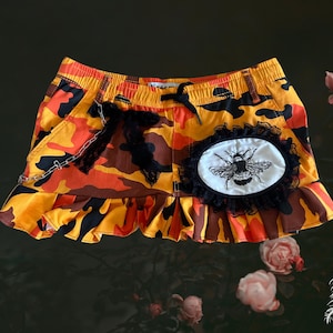 May include: An orange, yellow, and brown camouflage print skirt with a ruffled hem, an elastic waistband with a black drawstring, a silver chain detail, and a circular white patch featuring a black embroidered bee surrounded by black lace trim.