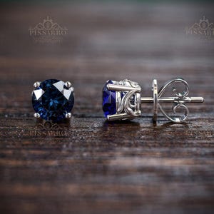 May include: A pair of stud earrings featuring round, deep blue gemstones set in silver-colored metal. The earrings are displayed on a dark wooden surface, showcasing their sparkle and intricate design. The brand name "Pissarro" is subtly visible.
