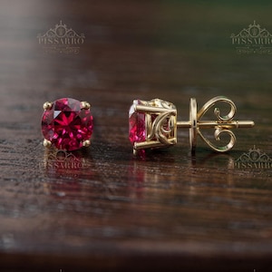 May include: A pair of gold stud earrings featuring round, deep red gemstones. The earrings are set in a classic four-prong setting, with a post and butterfly backing. The earrings are displayed on a dark wooden surface.