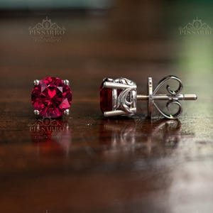 Puede incluir: Un par de pendientes de botón con gemas redondas de color rojo intenso engastadas en metal plateado. Los pendientes se exhiben sobre una superficie de madera oscura, mostrando los intrincados detalles del engaste y el color vibrante de las piedras.