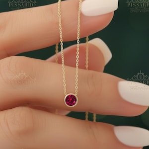 May include: A delicate gold chain necklace featuring a single round, bezel-set deep red gemstone pendant. The necklace is held by fingers with light-colored manicured nails. The text PISSARRO is visible in the background in multiple locations.