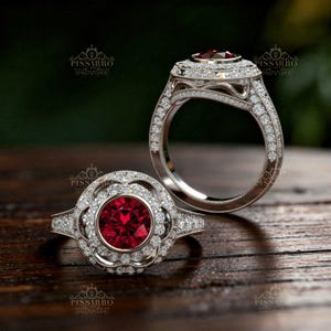 May include: Two silver rings with a large, round, red gemstone center. The rings are encrusted with small, clear gemstones. The rings are set on a wooden surface. The rings have a floral design around the center stone.