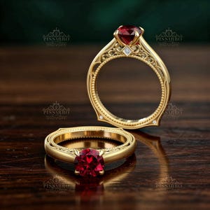 May include: Two gold rings with ornate detailing and a central red gemstone. The rings have a vintage style with intricate patterns and small accent stones. The rings are displayed on a dark wooden surface.