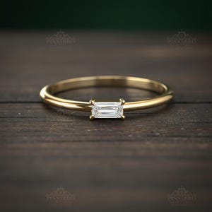 May include: A delicate gold ring featuring a rectangular, clear gemstone. The ring has a thin band and a simple, elegant design. The gemstone is set in a four-prong setting, showcasing its shape and clarity. The ring is displayed on a dark wooden surface.