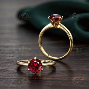 May include: A gold ring featuring a round, faceted deep red gemstone set in a six-prong mounting. The ring is displayed in two positions, one lying flat and one standing upright, with the PISSARRO logo visible in the background and foreground.