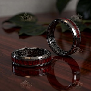 May include: Two dark metal rings featuring a central band of square-cut red stones. The outer edges of the rings have a beaded texture, while the inner surfaces display intricate, dark, engraved patterns. The rings are positioned on a dark surface.