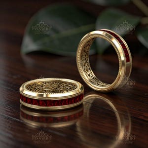 May include: Two gold rings featuring a channel-set row of square-cut red gemstones. The bands have a beaded edge and an intricate, openwork filigree pattern on the inner surface. The PISSARRO logo is visible on the image surface.