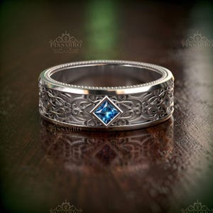 May include: A silver ring with an intricate floral design and a square, blue gemstone at the center. The ring has a detailed pattern around the band and a textured inner edge. The ring is displayed on a dark wooden surface.