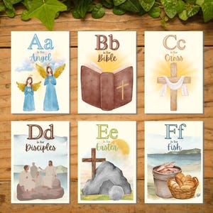 May include: A set of six watercolor art prints featuring religious themes. Each print displays a letter of the alphabet with a corresponding word: Angel, Bible, Cross, Disciples, Easter, and Fish. The prints have a cream background and are arranged on a wooden surface.