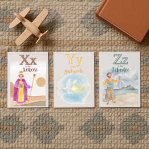 May include: Three illustrated alphabet cards with the letters X, Y, and Z. The X card features Xerxes, the Y card features Yahweh, and the Z card features Zebedee. A wooden toy airplane and a brown book are also in the image.