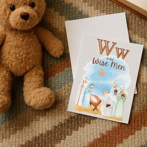 May include: A greeting card with the text "W is for Wise Men" and an illustration of the Nativity scene. The card is light gray with a watercolor-style image of the three wise men, Mary, Joseph, and baby Jesus. A brown teddy bear is in the background.