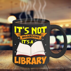 May include: Black coffee mug with a white open book and the text "It's not hoarding it's a library" in yellow.