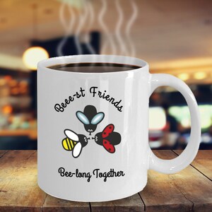 May include: White ceramic coffee mug with a black handle. The mug has a black and white illustration of a bee, a fly, and a ladybug with the text "Bee-st Friends Bee-long Together".
