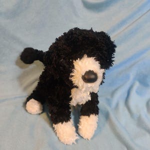 May include: A plush dog toy featuring a black and white shaggy texture. The stuffed animal has a black body with white patches on the chest, face, and paws, along with a smooth black plastic nose and dark eyes, standing in a four-legged pose.