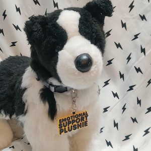 Może przedstawiać: Pluszowy pies w kolorze czarno-białym, z szarą obrożą i żółtą zawieszką z napisem "EMOTIONAL SUPPORT PLUSHIE". Tło to biała tkanina z czarnymi wzorami błyskawic.