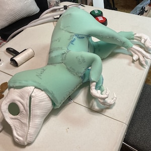 Peut inclure: Un patron de costume de dinosaure en mousse verte avec des marques manuscrites pour la couture. Le patron est disposé sur une table blanche avec une tête de dinosaure en mousse blanche.